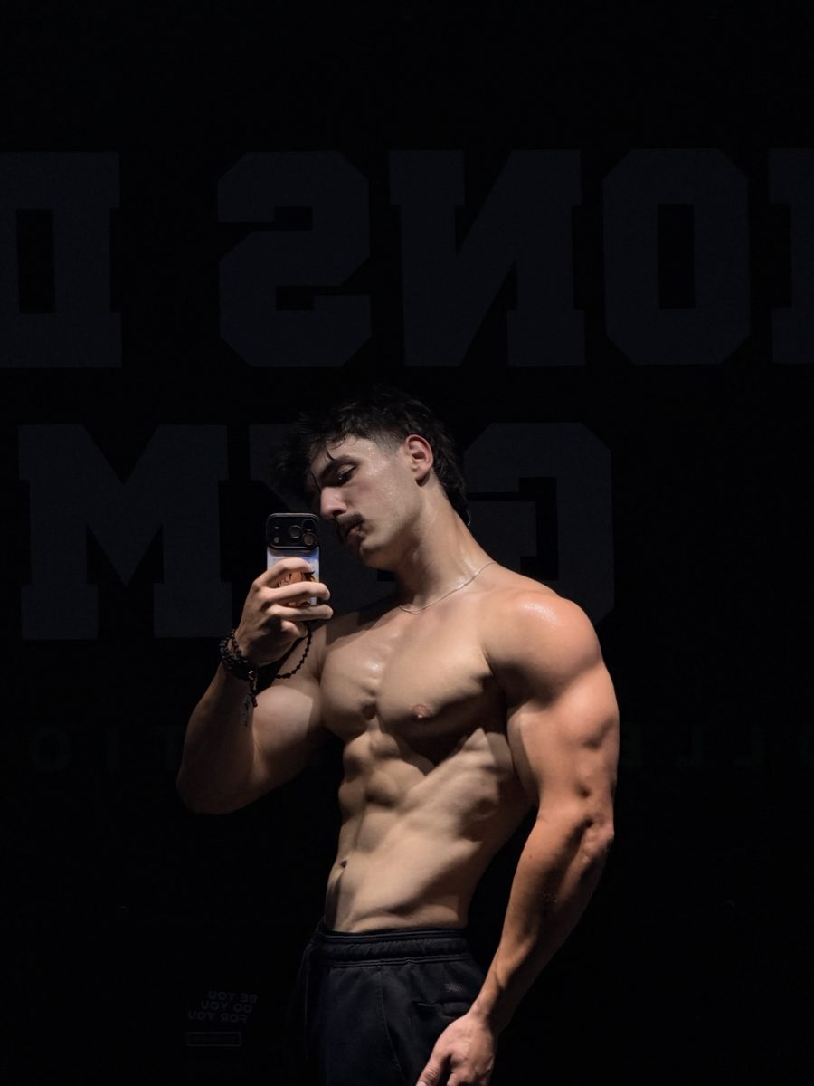 Alan flexing shirtless in a gym mirror